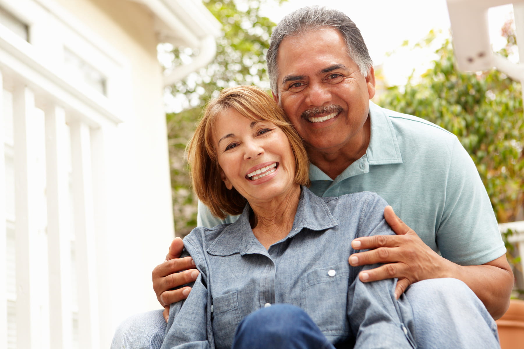 tooth implant restorations from Firm Foundation Dental in Prosper TX Smiling older couple poses together outdoors. The woman sits in front, wearing a denim shirt, while the man in a light blue polo stands behind her with his arms around her shoulders. Both look happy and relaxed.
