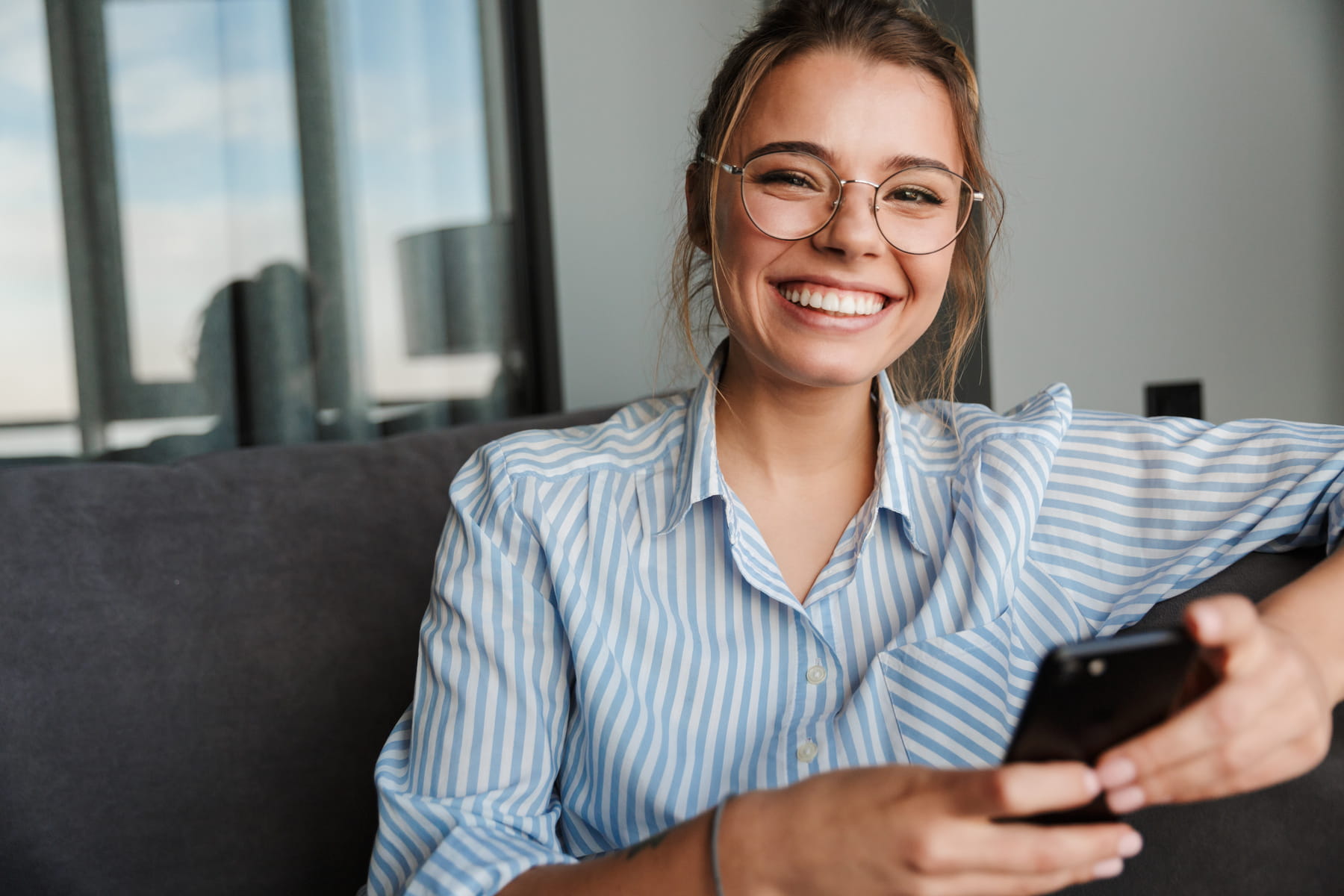 smile makeovers from Firm Foundation Dental in Prosper TX Smiling young woman wearing glasses and a striped shirt sits on a couch, holding a smartphone in one hand, with large windows in the background.
